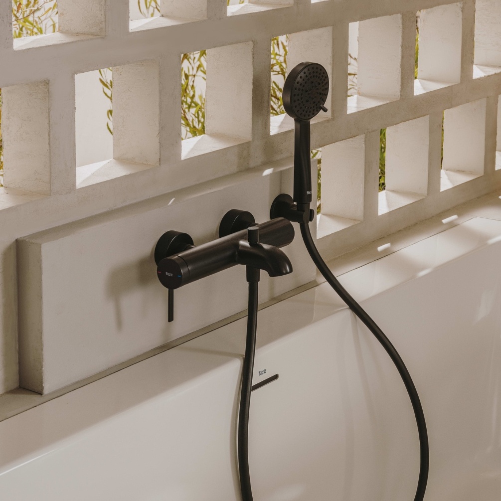 Lifestyle image of the Roca Ona Matt Black Wall Mounted Bath Shower Mixer mounted on a bathroom wall, above a bathtub and attached to a handset shower